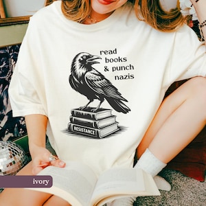 Op de afbeelding: Ivoor kleurig T-shirt met een zwarte raven illustratie op een stapel boeken. De tekst luidt "read books & punch nazis" boven de raaf en "RESISTANCE" op het onderste boek. Een casual kledingstuk.