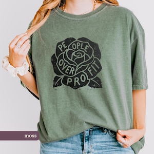 May include: Moss green t-shirt with a black rose graphic. The rose design features the words "People Over Profit" in a circular arrangement around the flower. The shirt is a relaxed fit and has short sleeves.