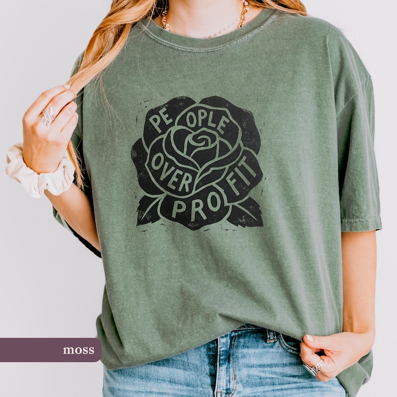 May include: Moss green t-shirt with a black rose graphic. The rose design features the words "People Over Profit" in a circular arrangement around the flower. The shirt is a relaxed fit and has short sleeves.