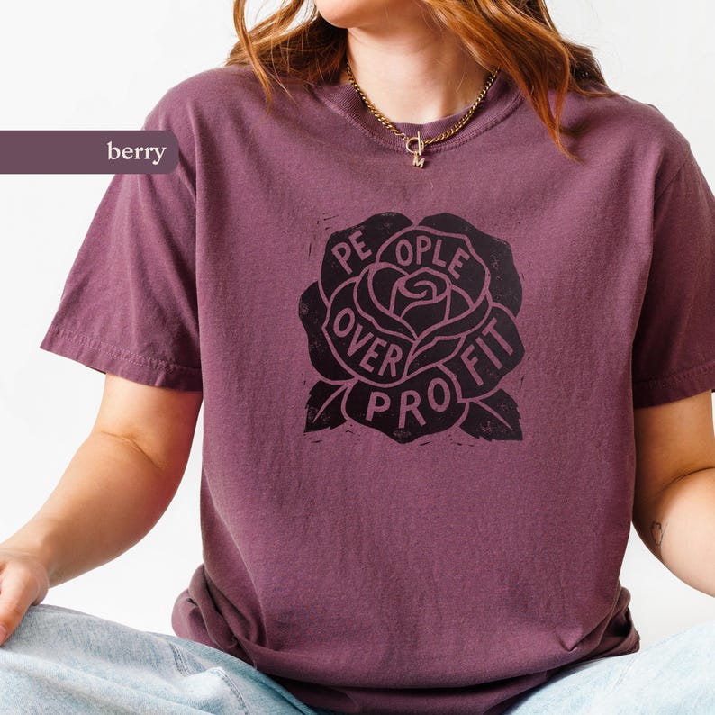 May include: Berry-colored t-shirt with a black rose graphic. The rose design features the words "People Over Profit" in a circular arrangement around the flower. The shirt has short sleeves and a crew neck.