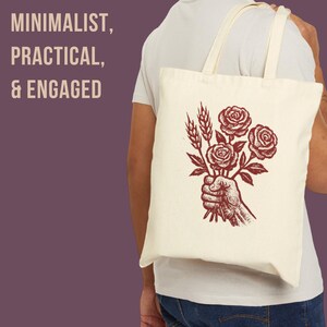 May include: A cream tote bag featuring a red illustration of a hand holding roses and wheat. The bag has the words "MINIMALIST, PRACTICAL, & ENGAGED" printed on the left side. The bag has two cream-colored handles.