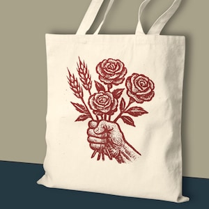 May include: Cream tote bag with a red illustration of a hand holding roses and wheat. The design is vintage-inspired. The bag has two long handles. The overall style is rustic and artistic.