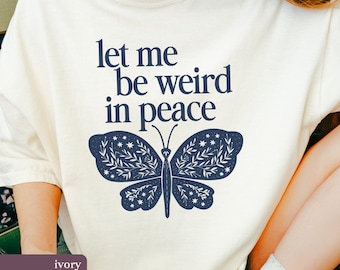 May include: Ivory t-shirt with the navy blue text "let me be weird in peace" and a navy blue butterfly design. The butterfly has leaf and star patterns. A casual wear item.