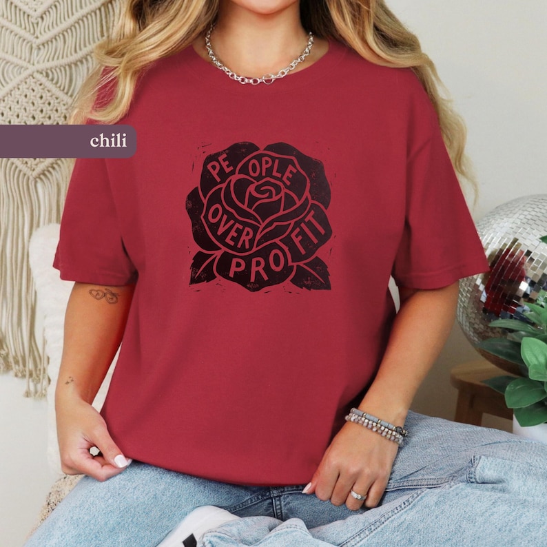 May include: Chili-colored t-shirt featuring a black rose graphic with the words "PEOPLE OVER PROFIT" in a circular design. The shirt has short sleeves and a crew neck. The word "chili" is printed on a rectangular label.