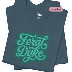 May include: Deep teal t-shirt with the words "Feral Dyke" in a teal script font with a dotted border. The shirt also features the text "hope RIOT" and "Heart-led rage..." in white. The shirt is folded.