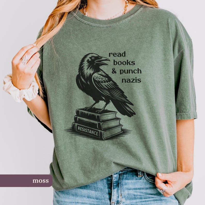 Op de afbeelding: Mosgroen T-shirt met een zwarte afbeelding van een raaf op een stapel boeken. De tekst luidt "read books & punch nazis" boven de raaf. Het onderste boek zegt "RESISTANCE".