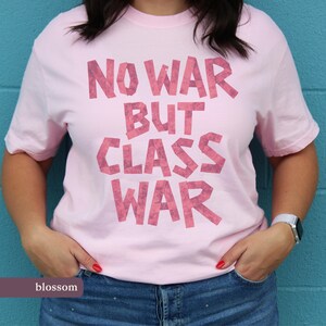 May include: A pale pink t-shirt featuring the slogan "NO WAR BUT CLASS WAR" in a textured, pink font. The short-sleeved shirt is made of a soft material. The word "blossom" is at the bottom of the image.