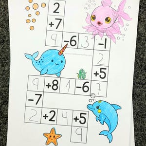 May include: A stack of white papers featuring a colorful, hand-drawn math worksheet. The worksheet includes addition and subtraction problems with cartoon sea creatures like a pink fish, blue dolphin, and narwhal. The website www.rainbowmaths.com is visible.