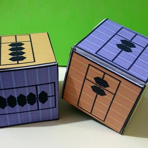 May include: Two colorful cube-shaped boxes with abacus-style designs. One box has a tan top and orange sides, the other has a purple top and sides. Each side features a black geometric pattern.