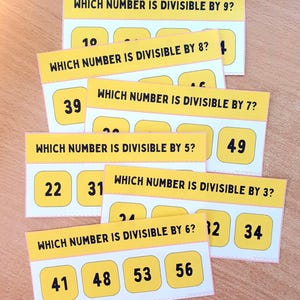 Op de afbeelding: Gele flashcards met de vragen "Welk getal is deelbaar door 9?", "Welk getal is deelbaar door 8?", "Welk getal is deelbaar door 7?", "Welk getal is deelbaar door 5?", "Welk getal is deelbaar door 3?" en "Welk getal is deelbaar door 6?" met getallen.