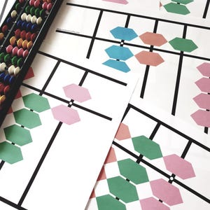 Könnte beinhalten: Ein schwarzer Abakus mit bunten Perlen liegt neben mehreren weißen Papierbögen. Die Papiere zeigen ein schwarzes Liniendesign mit grünen, rosa und blauen geometrischen Formen. Die Website www.rainbowmaths.com ist aufgedruckt.