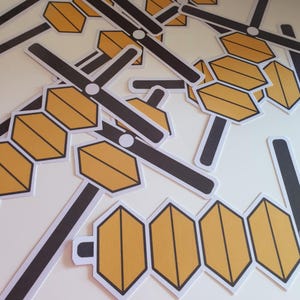 May include: A collection of paper cutouts featuring a yellow and black design. The cutouts have a hexagonal shape with a black border and a black bar extending from the hexagon. A white circle is at the intersection of the bar and the hexagon.