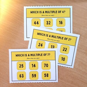 Op de afbeelding: Drie educatieve flashcards met gele randen en tekst. De kaarten vragen: "WELK IS EEN VEELVOUD VAN 4?", "WELK IS EEN VEELVOUD VAN 2?" en "WELK IS EEN VEELVOUD VAN 7?" Elke kaart heeft genummerde antwoordopties.