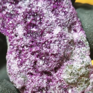 Puede incluir: Primer plano de una muestra mineral de color púrpura y gris con una textura rugosa y cristalina. El mineral tiene un aspecto brillante e iridiscente.
