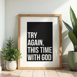May include: A rectangular wooden frame containing a white print with a black central rectangle. The text in white capital letters reads: TRY AGAIN, THIS TIME WITH GOD. The frame is positioned upright, displaying a clean and simple design aesthetic.