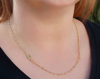 May include: A gold chain necklace with a small cross charm and a larger charm featuring the Virgin Mary. The necklace is worn by a person with long brown hair.