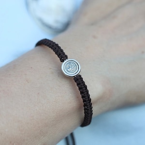May include: A brown braided bracelet with a silver charm featuring a detailed design. The bracelet is worn on a person's wrist.