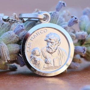 May include: Silver pendant featuring a depiction of Saint Mark, the Evangelist, holding a book and a lion. The pendant is on a chain and is resting on a sprig of lavender.
