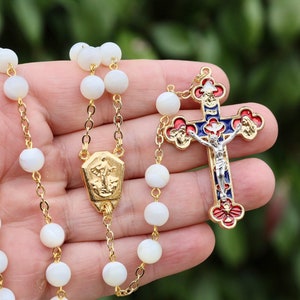 Puede incluir: Un rosario de perlas blancas con una cadena de oro y un crucifijo de oro y esmalte. El crucifijo presenta un diseño de esmalte azul, rojo y blanco.
