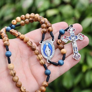 Könnte beinhalten: Ein Rosenkranz aus Holz mit blauen Perlen und einem silbernen Anhänger mit der Jungfrau Maria. Der Rosenkranz wird in einer Hand mit einer silbernen Uhr am Handgelenk gehalten. Der Hintergrund ist ein verschwommenes Bild von grünem Laub.