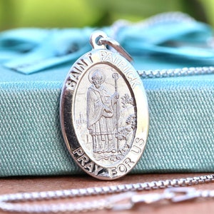May include: Silver oval pendant necklace with a Saint Patrick image and the words "Saint Patrick Pray For Us". The necklace is on a light blue gift box with a white ribbon.