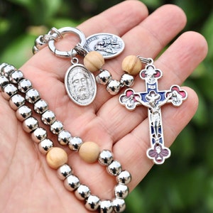 Op de afbeelding: Een religieuze sleutelhanger en rozenkrans set. De sleutelhanger heeft een zilverkleurige ovale hanger met een Jezus-afbeelding, een ronde hanger en een zilverkleurige ketting. De rozenkrans heeft zilveren kralen en een kruisvormige hanger met rode, blauwe en zilveren accenten.