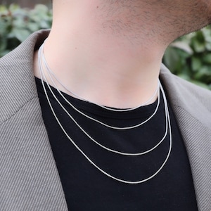 May include: A person wearing a black shirt and a gray blazer, showcasing a set of four silver chain necklaces of varying lengths. The necklaces are delicate and appear to be made of a fine, textured metal.