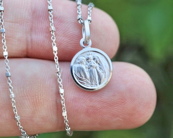 May include: A silver chain necklace with a round pendant featuring a detailed image of three figures. The chain is made of small, square links.