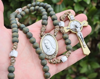 Può includere: Un rosario di legno verde con una medaglia d'argento e oro della Vergine Maria e un crocifisso d'argento e oro con una rappresentazione di Gesù Cristo. Il rosario è tenuto in una mano.