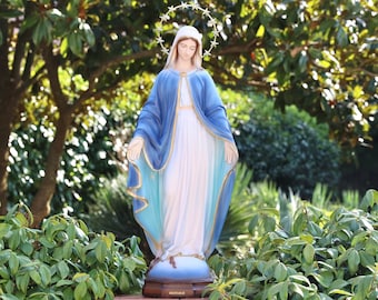 Könnte beinhalten: Eine weiße Statue der Jungfrau Maria mit einem blauen Gewand und einer goldenen Sternenkrone. Die Statue steht auf einem braunen Sockel mit der Aufschrift "MIRACULA". Die Statue steht vor einem grünen Busch.