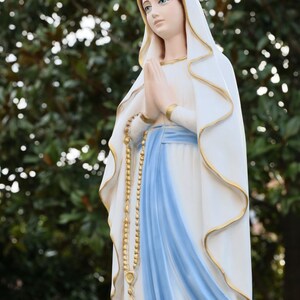 May include: A white statue of the Virgin Mary with a blue sash and a gold rosary. She is wearing a white veil and has her hands clasped in prayer.