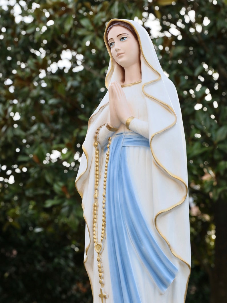 May include: A white statue of the Virgin Mary with a blue sash and a gold rosary. She is wearing a white veil and has her hands clasped in prayer.