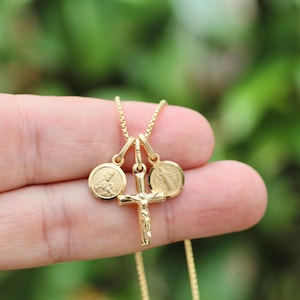 May include: Gold-colored necklace featuring a crucifix pendant and two round pendants. The crucifix has a detailed figure of Jesus. The round pendants have religious figures. The necklace is held against a blurred green background.