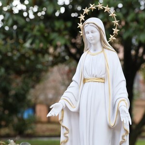 May include: White statue of the Virgin Mary with a gold star halo. The statue is wearing a white robe with gold trim and is standing with her arms outstretched.