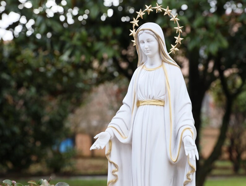 May include: White statue of the Virgin Mary with a gold star halo. The statue is wearing a white robe with gold trim and is standing with her arms outstretched.