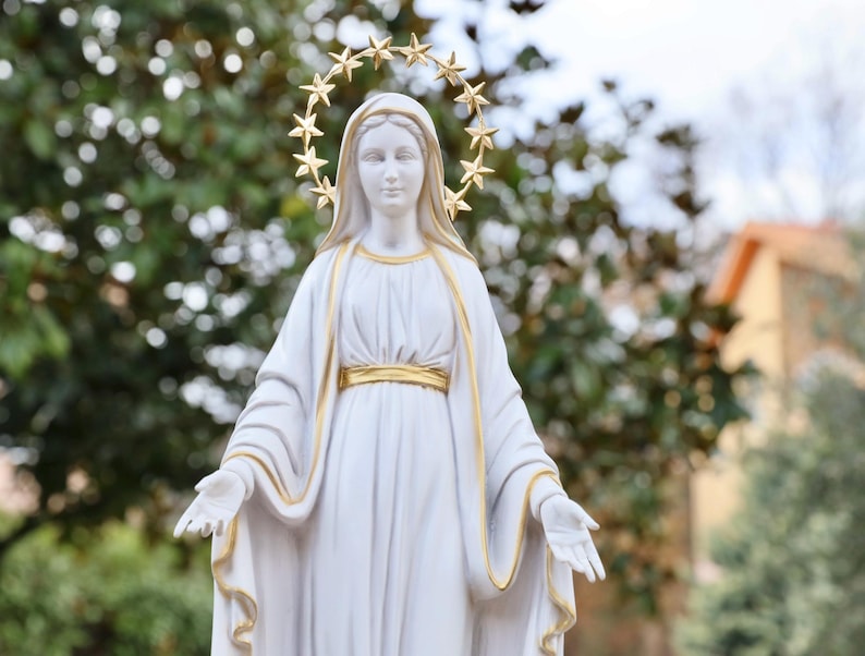 May include: White statue of the Virgin Mary with a gold star halo. The statue is wearing a white robe with gold trim and has her hands outstretched.