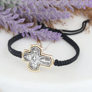 May include: A black braided bracelet featuring a silver and gold cross pendant. The cross has detailed religious imagery. The bracelet is adjustable with a sliding knot closure. The background includes purple flowers.