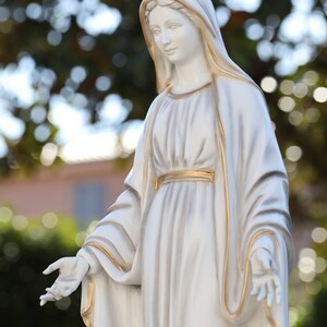 May include: White statue of a woman wearing a long, flowing robe with gold accents. The statue is standing with her arms outstretched.