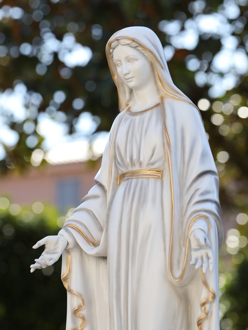 May include: White statue of a woman wearing a long, flowing robe with gold accents. The statue is standing with her arms outstretched.