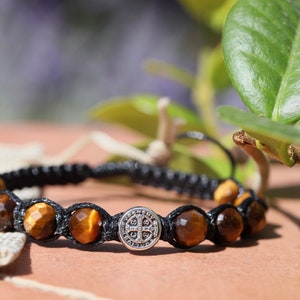 以下が含まれることがあります： 黒と茶色のタイガーアイビーズのブレスレット。シルバーの十字架チャーム付き。黒い紐で結ばれています。