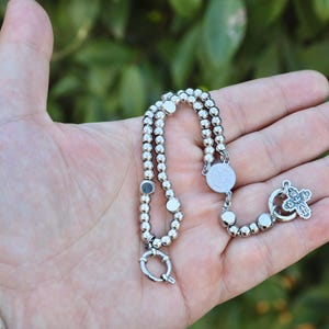 May include: A silver beaded bracelet with a silver cross charm and a silver clasp. The bracelet is on a hand.