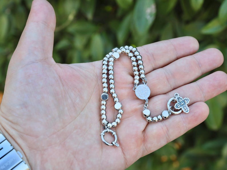 May include: A silver beaded bracelet with a silver cross charm and a silver clasp. The bracelet is on a hand.
