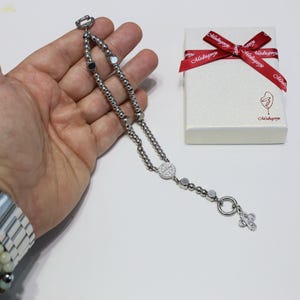 May include: A silver chain bracelet with a silver charm in the shape of a cross. The bracelet is on a hand and the charm is hanging down. The bracelet is in a white box with a red ribbon.