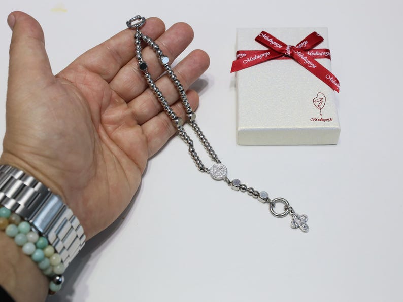 May include: A silver chain bracelet with a silver charm in the shape of a cross. The bracelet is on a hand and the charm is hanging down. The bracelet is in a white box with a red ribbon.