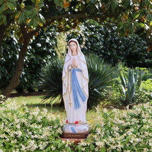 May include: A white statue of the Virgin Mary, wearing a blue robe and a crown of stars, stands in a garden setting. The statue is surrounded by green foliage and white flowers.