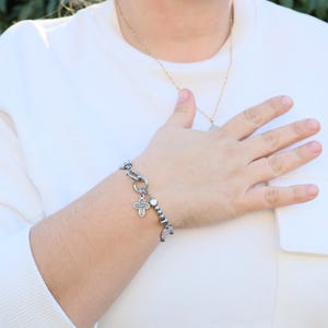 May include: A silver bracelet with a cross charm and small beads. The bracelet is on a person's wrist.