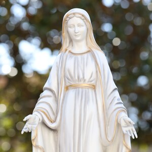 May include: White statue of a woman wearing a long white robe with gold trim. The statue is standing with her arms outstretched.