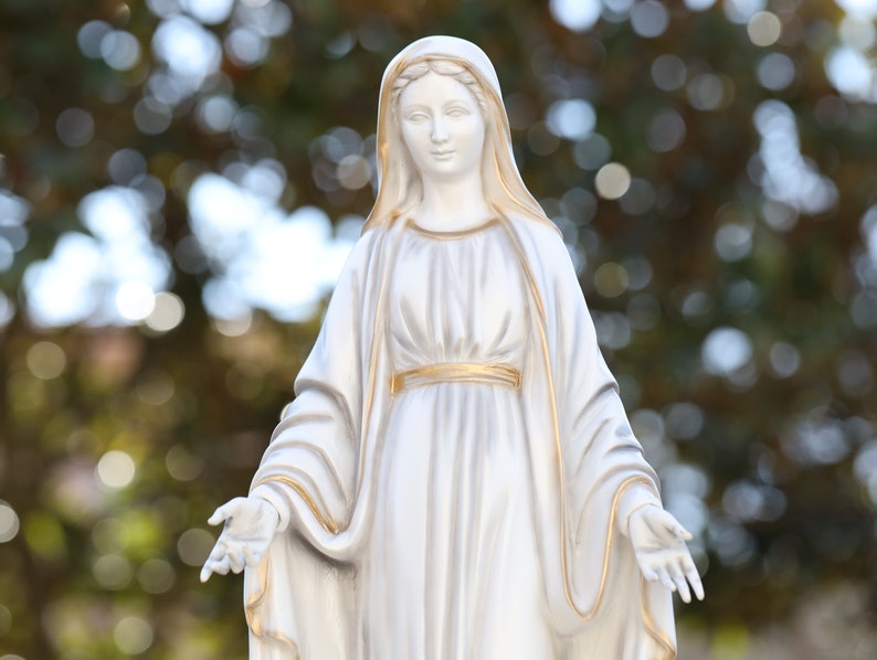 May include: White statue of a woman wearing a long white robe with gold trim. The statue is standing with her arms outstretched.