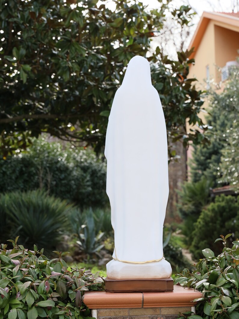 May include: White statue of a woman wearing a long robe, standing on a brick base in a garden setting.