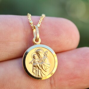 Pode incluir: Colar com pingente de ouro que apresenta um amuleto redondo com uma imagem detalhada de S&atilde;o Jos&eacute; segurando o menino Jesus. O amuleto tem uma borda elevada e um acabamento liso e polido.